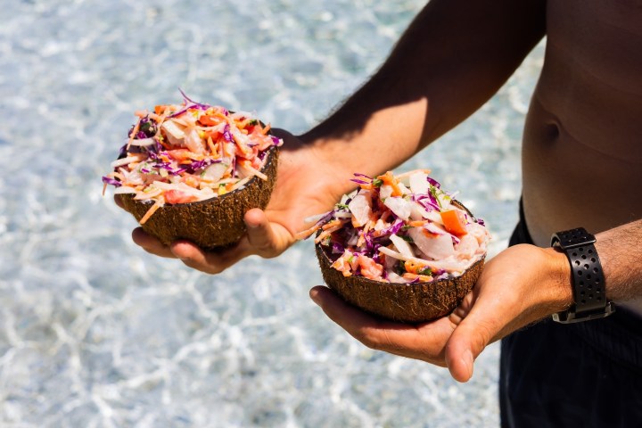 Person holding two coconut shells filled with colorful salad by the beach.
